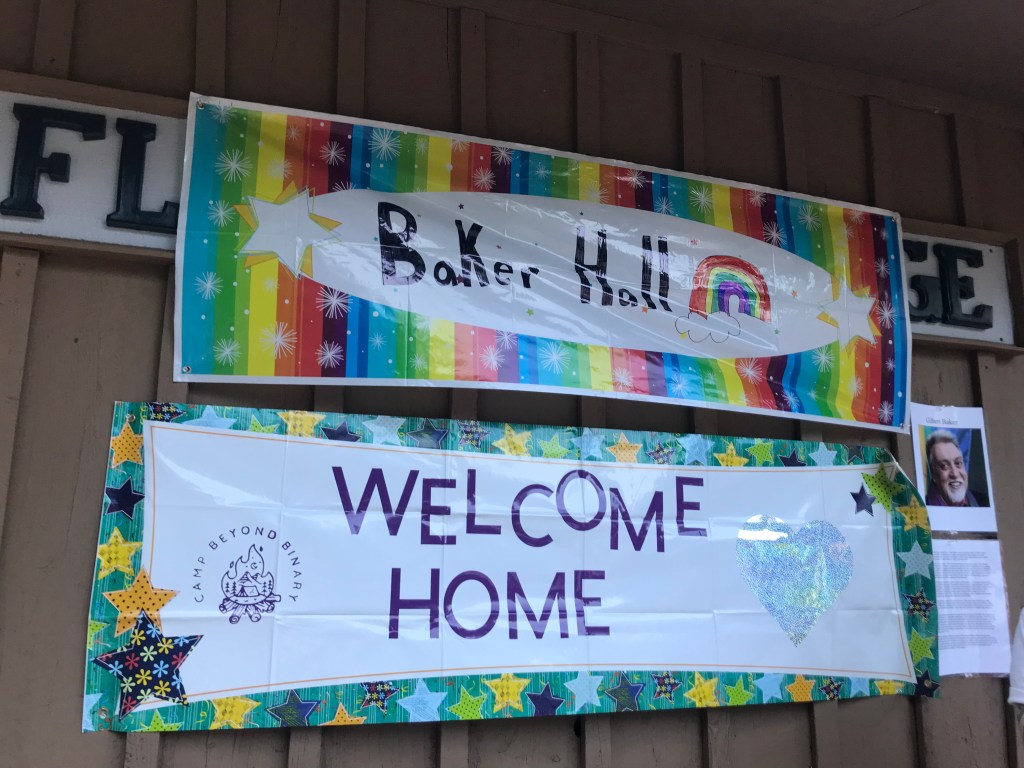 A rainbow banner that says "Baker Hall" with another banner below that says "Welcome Home." On the right side of the frame is a photo of Gilbert Baker who made the first Pride flag.